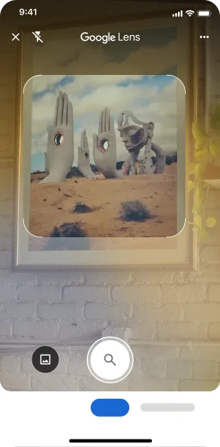 Sur un téléphone, l'appli Lentille Google est ouverte. Une notification indique à l'utilisateur qu'il peut toucher le bouton de l'obturateur pour rechercher une image de grandes sculptures de mains en pierre dans un désert.